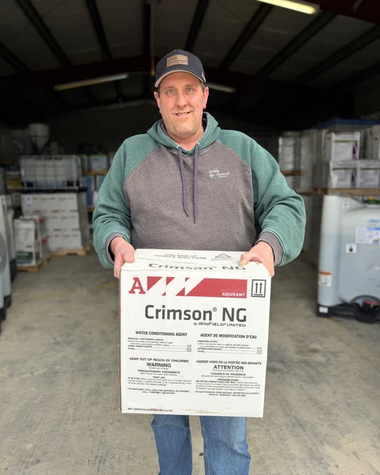 A person is standing in a warehouse holding a large box labeled "Crimson NG." They are wearing a cap and a two-tone hoodie. The background contains shelves with various boxes and containers.