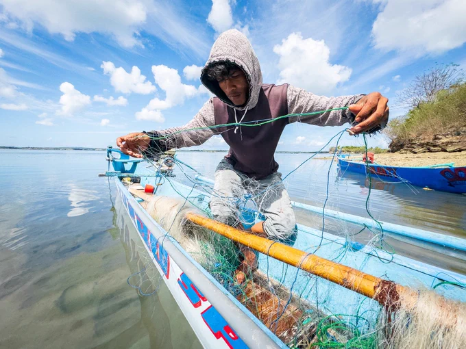 Back on the safety of the island beach, Pak Peri sits inside his small wooden boat, meticulously inspecting, cleaning, and repairing every inch of his large net. Despite the poor harvest, he prepares his gear with unwavering care right from the deck of his vessel for the next day's attempt.