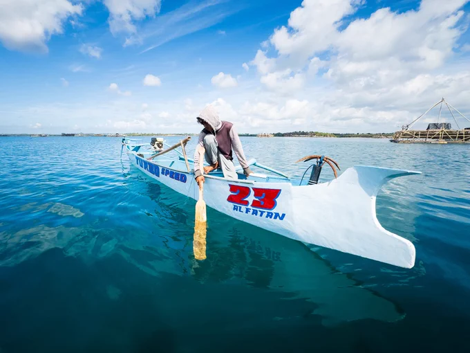 Leaning over the side of his boat, Pak Peri uses a long wooden oar to steady the craft while peering deep beneath the surface. The tension is clear; replacing or repairing these lost nets represents a devastating financial blow for a young father with no economic safety net.