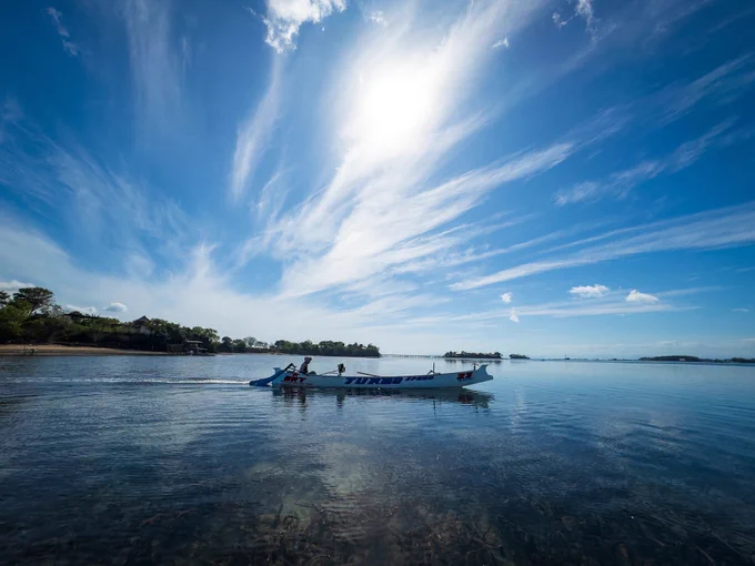 A wide documentary shot of 22-year-old fisherman Pak Peri standing at the stern of his traditional, narrow wooden motorised boat. He looks focused as he steers the craft away from the shores of his remote island off the Lombok coast, heading out into a vast, calm blue ocean to begin his daily hunt for crabs.