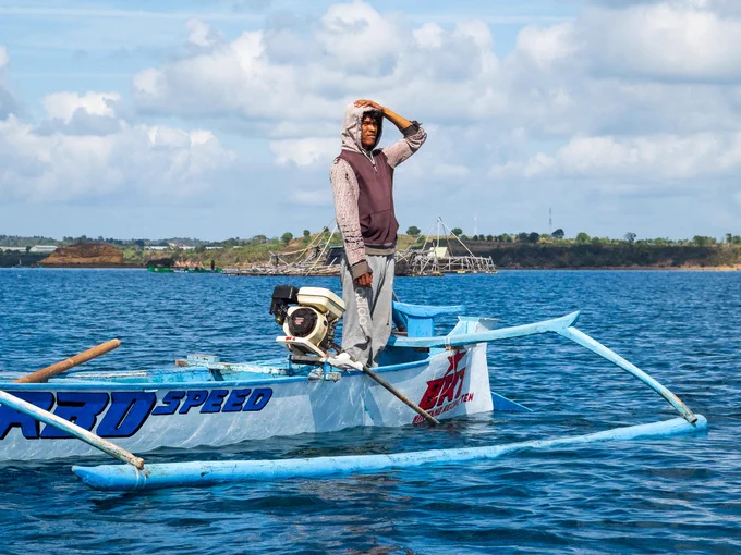 Pak Peri stands tall in his small wooden boat, shading his eyes from the harsh sun as he anxiously scans the open water. His fishing nets are missing—likely displaced by strong seasonal currents or severed by another vessel's engine—capturing the immediate, unpredictable vulnerability of artisanal fishing.