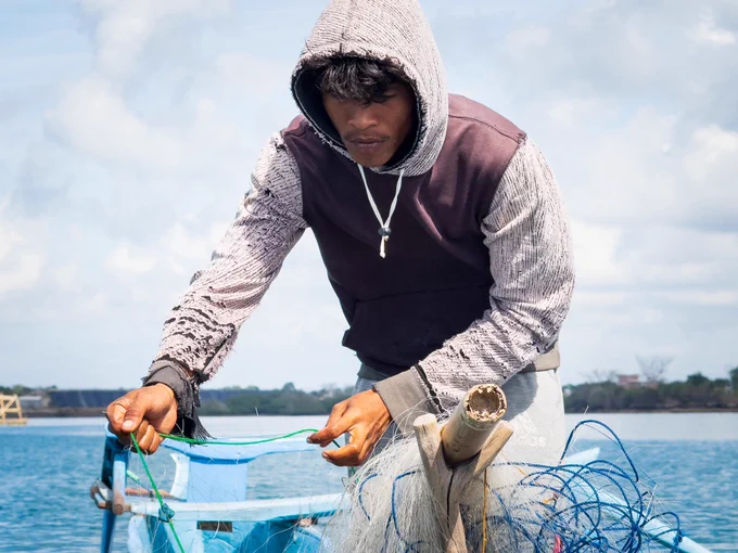 A gritty, close-up documentary shot of Pak Peri’s hands pulling the heavy, water-logged fishing net hand-over-hand into the boat. The physical strain highlights the raw, unmechanized labour required to sustain a family in this remote island community.