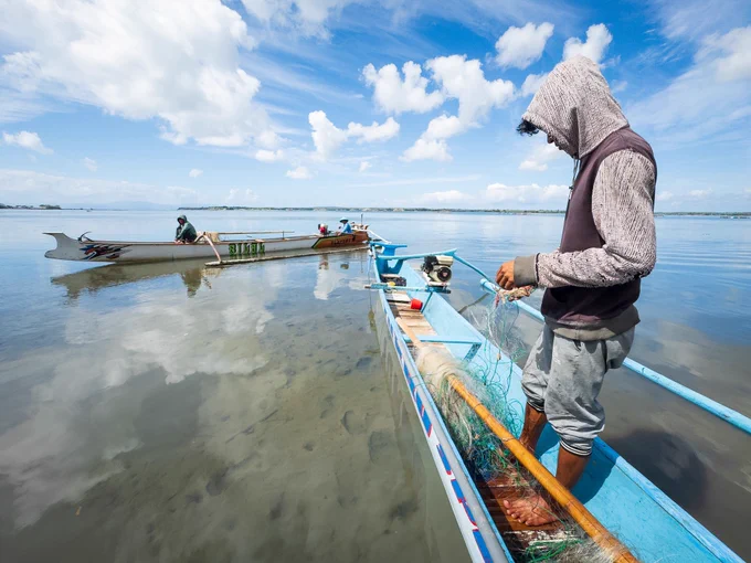 A wide narrative shot of Pak Peri working diligently on his nets on the shoreline, while another traditional island fisherman's boat pulls up onto the beach in the background—a reflection of an entire community bound to the seasonal struggles of the sea.
