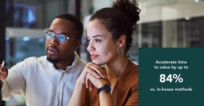 Two colleagues engaged in a focused discussion in a modern office setting, collaborating on a project. Overlay text "Accelerate time to value by up to 84% vs. in-house methods."