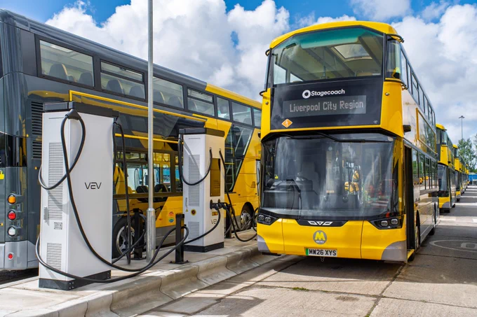 A fleet of bright yellow and grey electric double-decker buses parked at an outdoor charging depot, with the forefront bus plugged into a white VEV charging unit under a blue, cloudy sky.