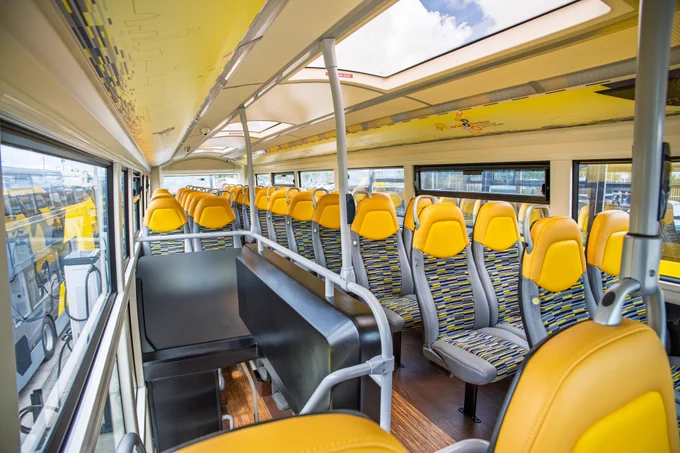 The modern interior of the upper deck of a bus, featuring rows of grey passenger seats with bright yellow headrests and patterned fabric, grey handrails, and a view of another yellow bus through the side window.