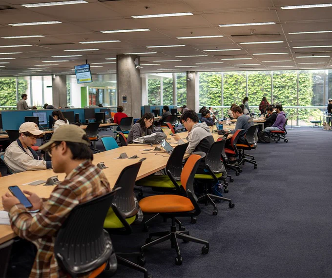The UCSD library filled with individuals sitting at long tables, studying or using laptops. 