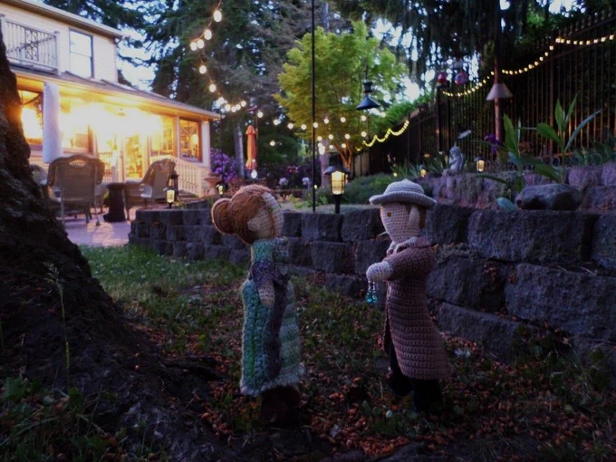 Crochet dolls of Mary Woodhull & Marshal Kahler, who is holding up the earrings. They are on the back lawn at twilight with retaining wall, house, trees & solar lights in background.