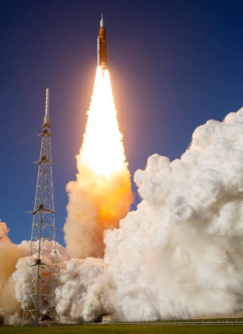 NASA's Artemis II mission lifts off atop the orange Space Launch System rocket from the launch pad at NASA's Kennedy Space Center. White, pillowy plumes fill the bottom half of the photo; a dark blue evening sky fills the top half. Credit: NASA/Keegan Barber