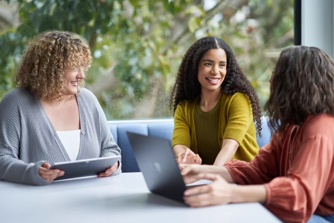 Three women sitting around each other at a table, smiling and collaborating on their tablet and computer 