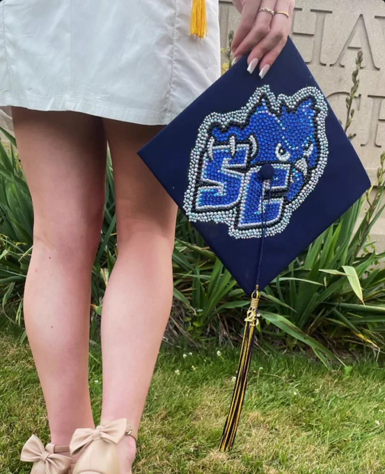 A person in a white outfit holds a decorated graduation cap featuring the SCSU Athletics logo. The cap is embellished with blue and silver sequins. Nearby are green plants and a stone wall.