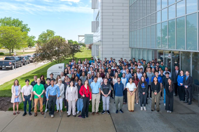 A large group of people wearing professional attire pose for a photo outside in front of NASA Glenn's Mission Integration Center building. A tree-lined road stretches away from them in the background and the sky is blue with a few white clouds. Credit: NASA/Jef Janis