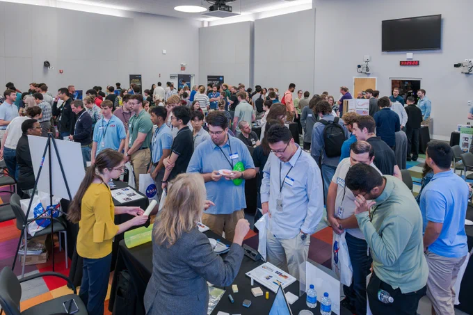 Visitors interact with NASA Glenn team members who are set up at tables throughout an indoor conference room space. Attendees wear nametags and lanyards, and several pop-up banners are set up behind the tables around the room. Credit: NASA/Jef Janis