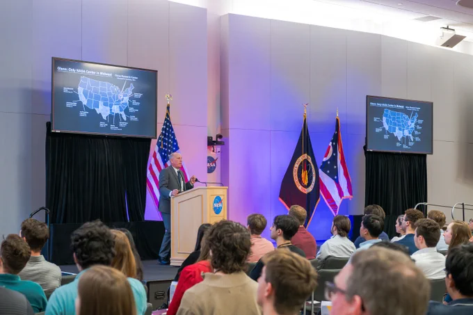 NASA Glenn's Larry Sivic speaks to a large group inside an indoor auditorium space. He stands at a tan podium, and behind him are NASA, Ohio, and American flags and a monitor that says, "Glenn: Only NASA Center in Midwest." Credit: NASA/Jef Janis