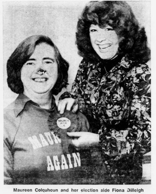 Black and white photograph of Maureen Colquhoun and her aide Fiona Dilleigh. Colquhoun is shown left, wearing a sweater reading 'Maureen Again.' She has a short dark bob. Next to her, with feathered hair, standing taller, is Dilleigh, who wears a floral patterned shirt and is pinning a rosette on Colquhoun, which readings 'Maureen again.'