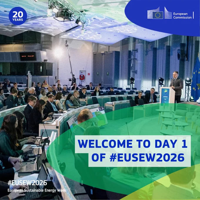 A photo of a modern conference hall filled with seated attendees using laptops and taking notes during a formal event. At the front right, a man stands at a lectern speaking, with EU branding visible nearby. The European Commission logo appears in the top right corner, and a '20 YEARS' badge is shown in the top left. Overlaid graphic text reads 'WELCOME TO DAY 1 OF #EUSEW2026,' with additional branding at the bottom left stating '#EUSEW2026 European Sustainable Energy Week.' 