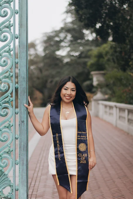 Young woman in a white dress and Berkeley graduation stole stands smiling near an ornate green gate. 