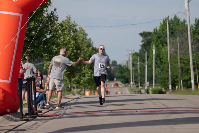 A trooper participates in the 6th Annual Rick Moyer Legacy Foundation Pump n Run/5K. He is approaching the finish line and is giving a high five to someone along the course.