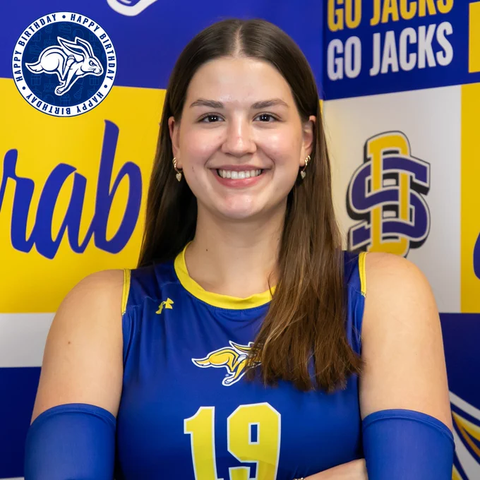 Women's volleyball player in a blue jersey and blue arm sleeves stands with arms crossed on a branded SDSU background. A blue and white, circular "happy birthday" graphic appears in the top left corner with a Jackrabbit logo in the middle. 