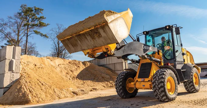 A John Deere loader is scooping a large pile of sand at a construction site. The machine's bucket is raised, and there are large concrete blocks in the background. Trees and a clear blue sky are visible.