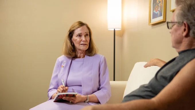 Dr. Leslie Robbins, dean of the UTEP College of Nursing, in a light purple outfit is sitting on a sofa in a softly lit room, writing in a notebook. Facing her is another individual with short hair and glasses, casually dressed, sitting on the same sofa. A floor lamp and two framed pictures are visible in the background.