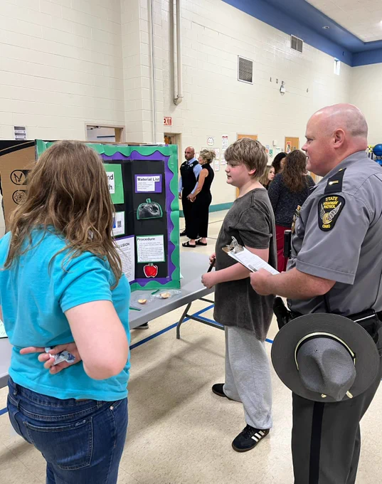 Lt. Morrison judging a project at the science fair. Two students stand next to their project. 