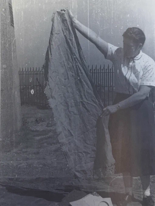 Historical black-and-white photograph of Sally Wollaston standing outdoors at Blue Hill Observatory holding up a large dresser scarf recovered after the Worcester tornado of June 9, 1953. The Observatory's fencing can be seen in the background, while the scarf, carried more than 25 miles by the storm, fills much of the frame.