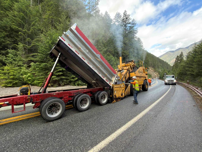A truck dumps hot asphalt into a large yellow paver along a mountainous stretch of State Route 20/North Cascades Highway. Workers and other equipment are in the area.