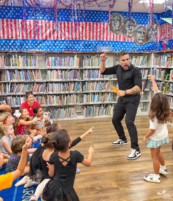 A man performs for a group of young children in a library's 'Children's Area'. He wears black, with an animated expression, holding an orange object, as children sit on the floor, pointing and laughing. The room has American flag decor with Mount Rushmore heads.