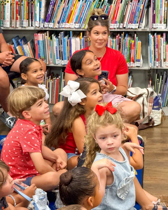 A group of children on a blue rug in a library. Bookshelves filled with colorful books form the background. Some children look up smiling. 