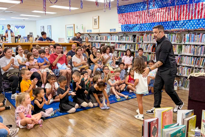 A man in a black shirt interacts with a child at a library event. Children sit on a blue mat, clapping, while adults watch. 
