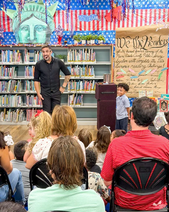 A magician in a black vest is standing in front of kids & adults in a library. A young boy stands smiling near him. 