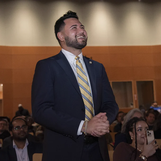 A person in a suit and yellow-striped tie is smiling and standing in an auditorium. The audience is in the background, and someone is taking a photo with a smartphone.