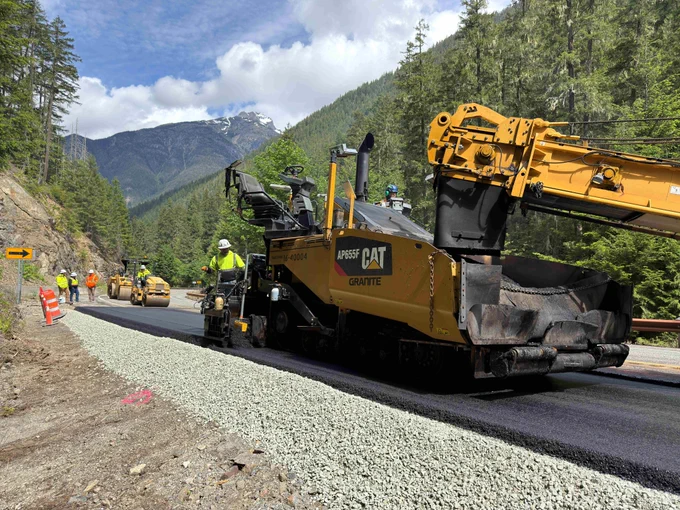 A large yellow paver lays down fresh asphalt in a mountainous area of State Route 20/North Cascades Highway. Other workers and equipment is seen in the background.