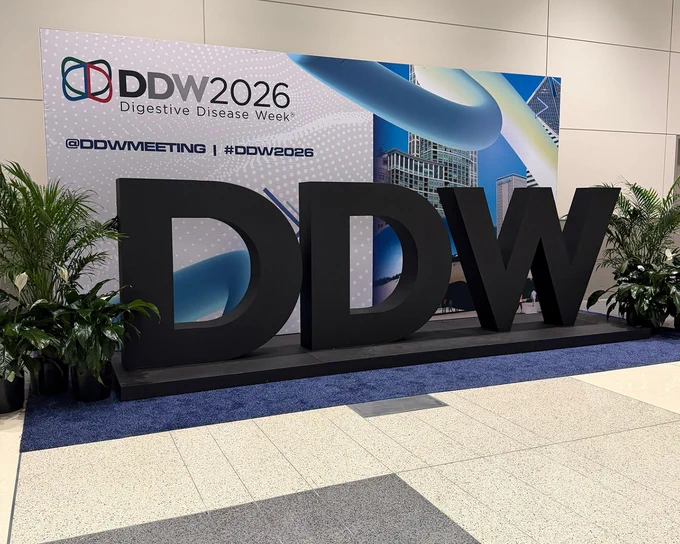 Large black 3D letters DDW in front of a DDW2026 backdrop for Digestive Disease Week, flanked by potted plants.