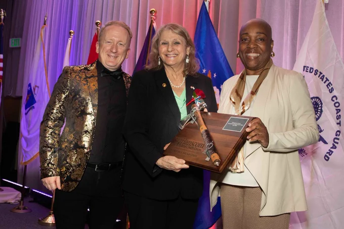 Three individuals stand together at an awards ceremony, with one holding a wooden plaque and ceremonial staff, surrounded by flags in the background.