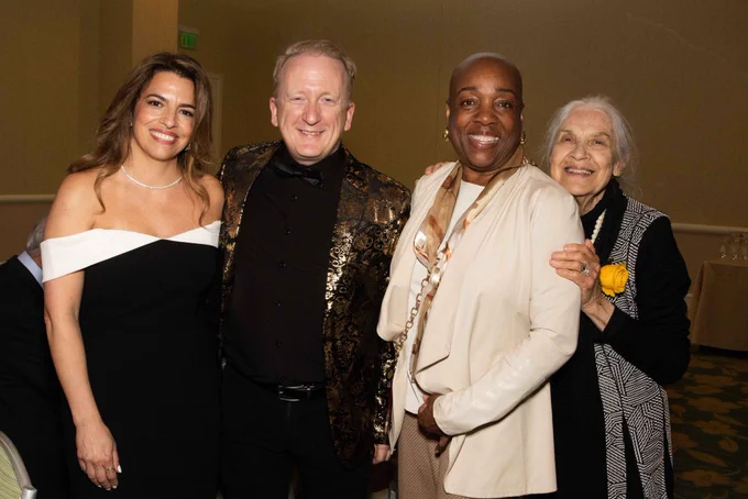 Four elegantly dressed individuals posing together at an indoor event with a carpeted floor and beige walls.