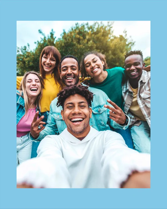 A group of six people smiling and taking a selfie outdoors with greenery in the background. They are casually dressed and appear joyful.

