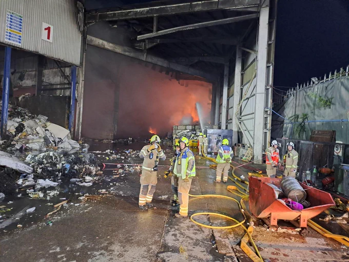 A group of firefighters stand in front of a fire at a recycling centre, which is contained to one area at the back of the picture.