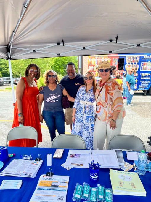 A group of five people smile together under a canopy tent behind a blue table filled with "Safer Stronger Together" community resource materials on District Heights Day.