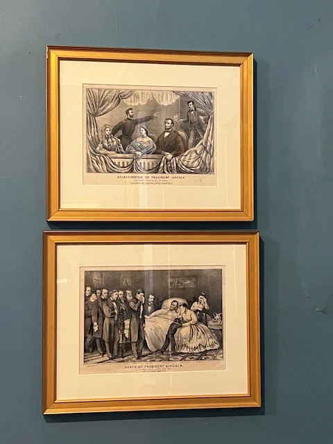 Two framed illustrations on a blue wall. The top illustration depicts the assassination of President Lincoln, featuring several figures around a bed. The bottom illustration shows a group gathered around a bed, titled "Death of President Lincoln.