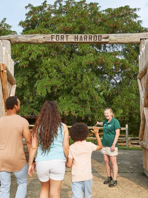 Family entering Fort Harrob, greeted by a person in green attire under a wooden archway with trees in the background.