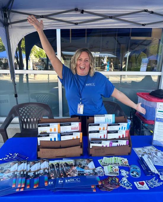 An RCC employee shows off all the take-away items at an RCC booth during a public event in Medford. She smiles in a blue RCC shirt with her hands in the air, showing enthusiasm.