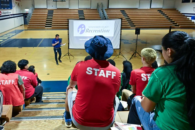 Staff members in red shirts seated on gym bleachers listening to a speaker presenting in front of a recreation department screen.