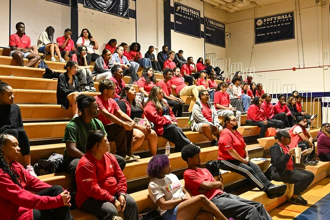 A diverse group of people seated on wooden bleachers inside a gymnasium, attentively watching a MoCoRec staff training session.