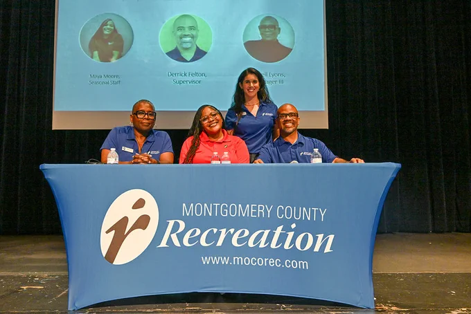 Four individuals seated and standing behind a Montgomery County Recreation table on a stage with a projected presentation behind them.