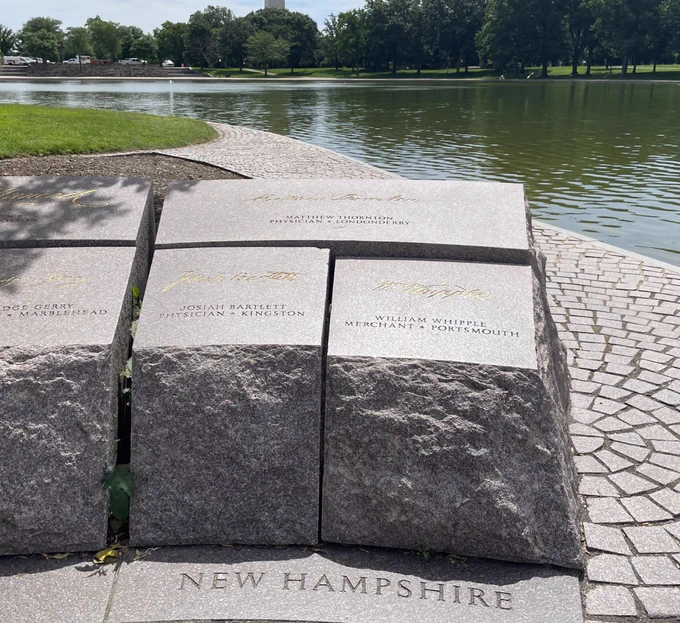 Three reddish gray stone blocks with names etched into the tops and gold signatures on them and the words "New Hampshire" written at the base stand as a section of a low memorial wall with a calm pond and a park in the background.