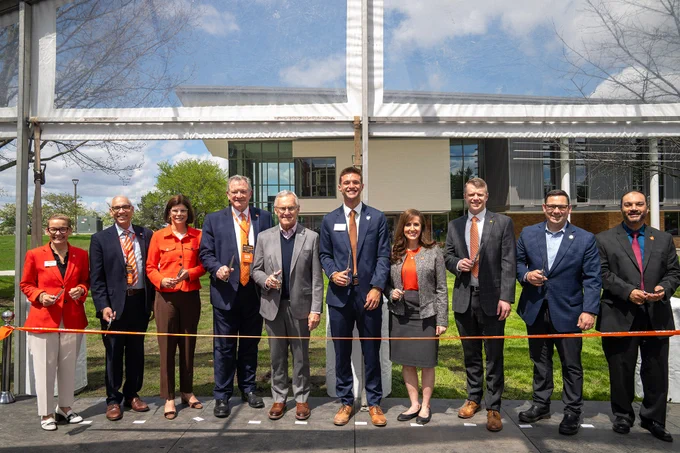 A group of people standing outdoors at a ribbon-cutting ceremony. They are holding scissors and wearing business attire. Behind them is a building with large windows and a grassy lawn. A ribbon spans in front of the group, ready to be cut.