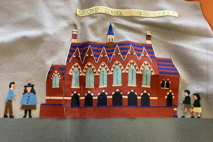 A fuzzy felt rendering of the exterior frontage of a school. Banner at the top of the artistic work reads Second School 1870 – 1957. The school has chimneys at either end of the roof and a bell tower in the middle. A line of vaulted windows running across the middle of the building. A series of archways below the windows. An entrance on the far right. Figures of young people at either end of the building. 
