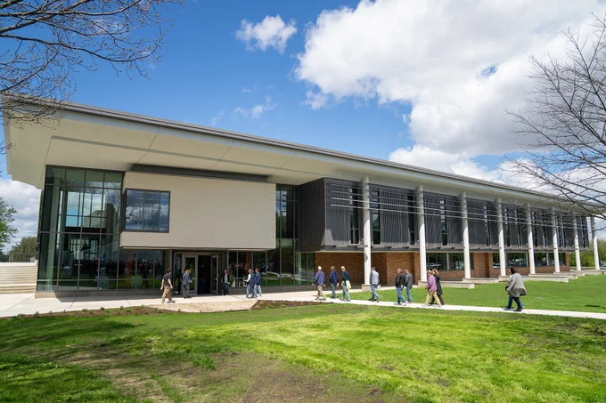 A group of people walk towards a modern, glass-fronted building on a bright, sunny day. The building is situated on a well-maintained grassy area with a few trees, under a partly cloudy sky.